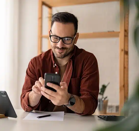 A man is smiling, wearing casual clothes, and using a modern smartphone in his home office