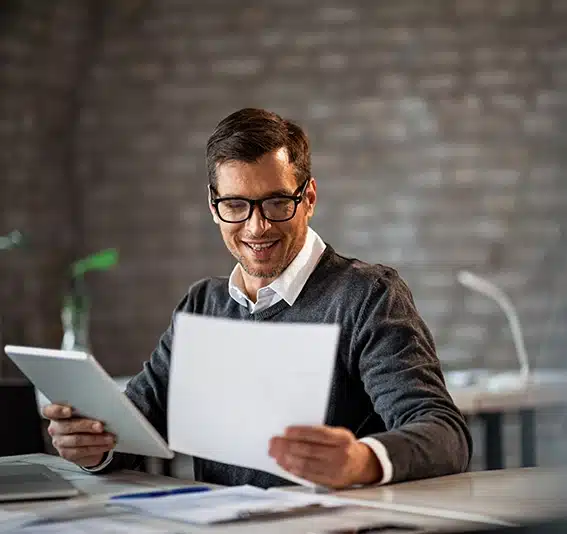 A man with glasses at work is holding a document and using a digital tablet