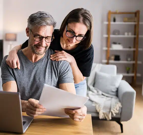 A couple with glasses holding a document in front of a laptop screen