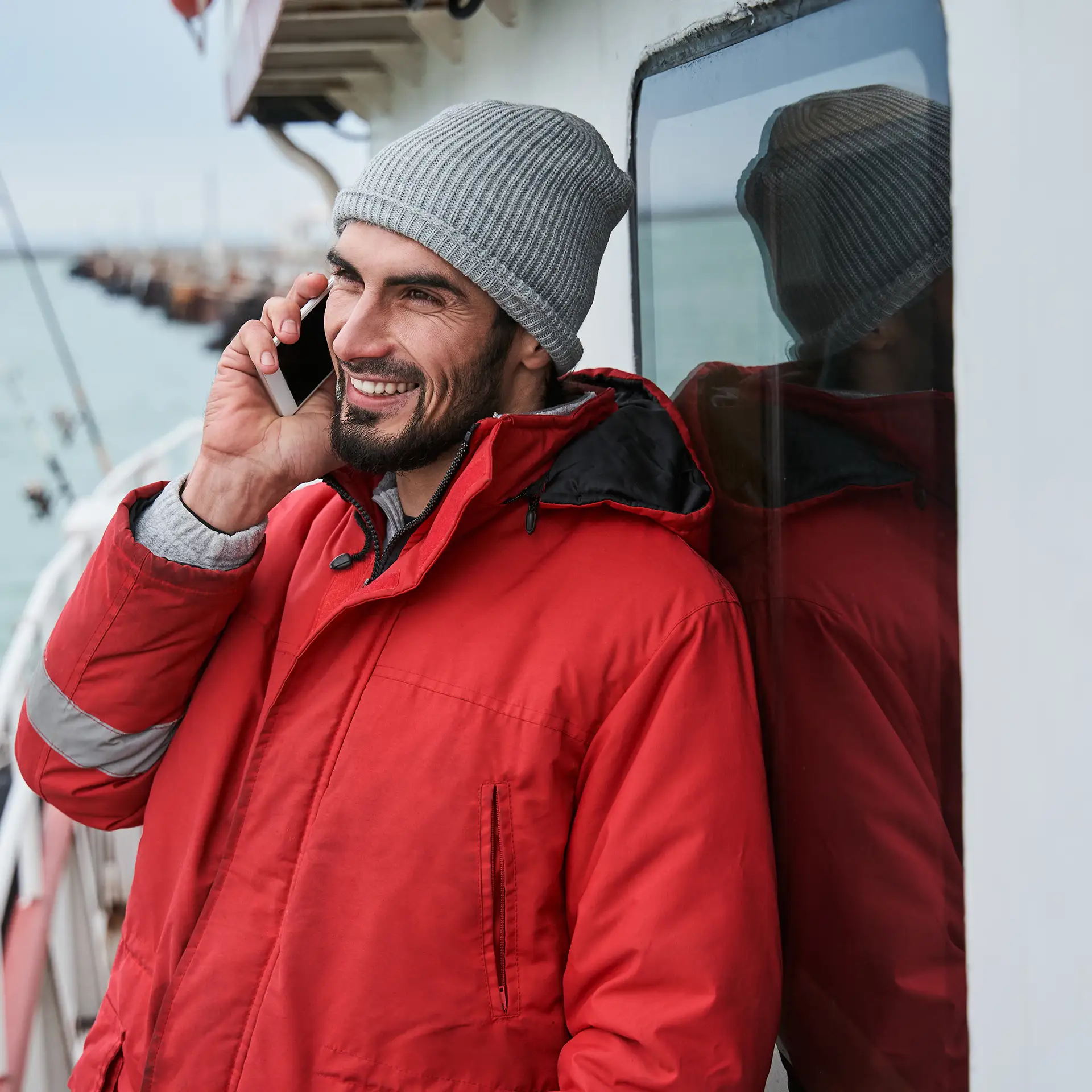Side view of a young, unshaven man in a warm jacket standing on the boat deck and using his smartphone