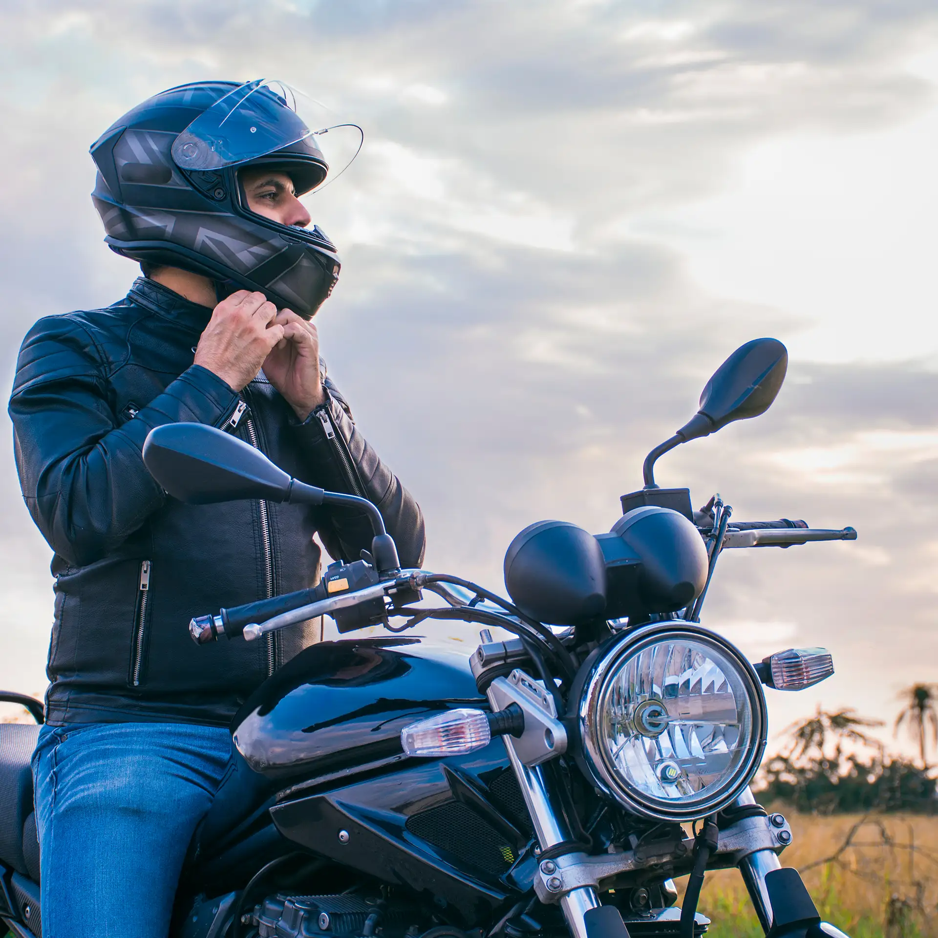 Man sitting on a motorcycle, wearing jeans and a black jacket, fastening his helmet with a landscape in the background