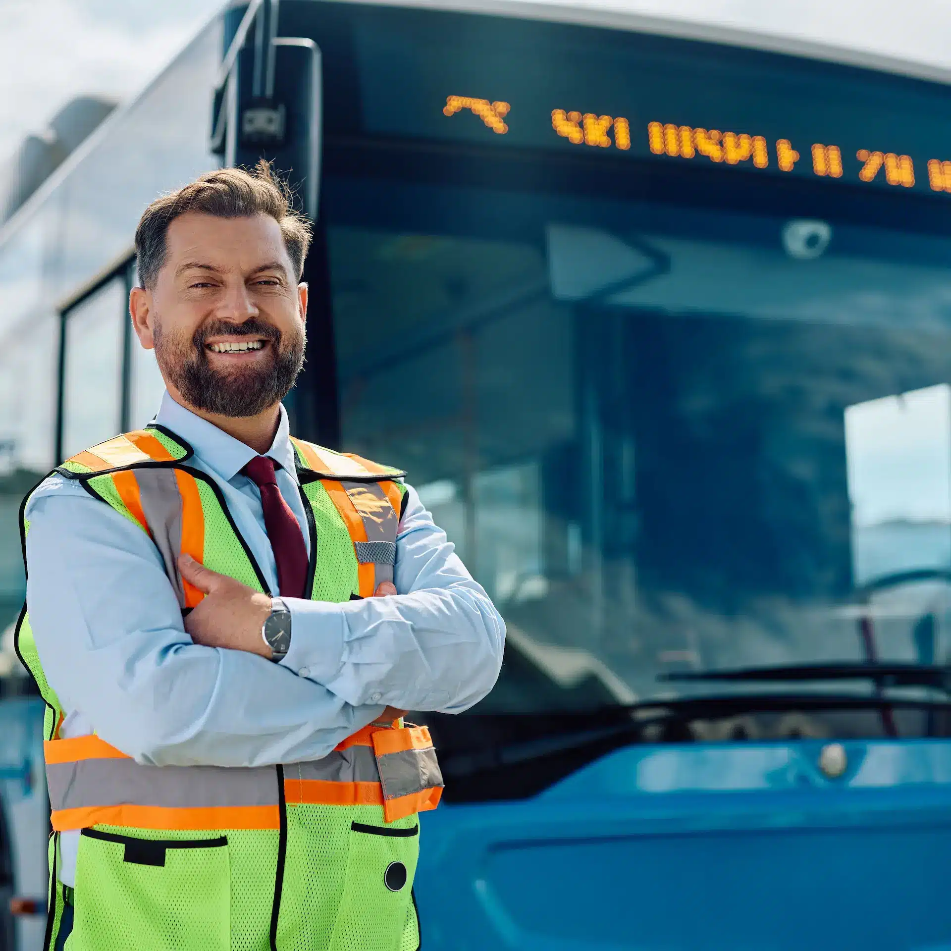 A bus driver proudly standing next to his bus wearing a high visibility vest with his arms crossed smiling