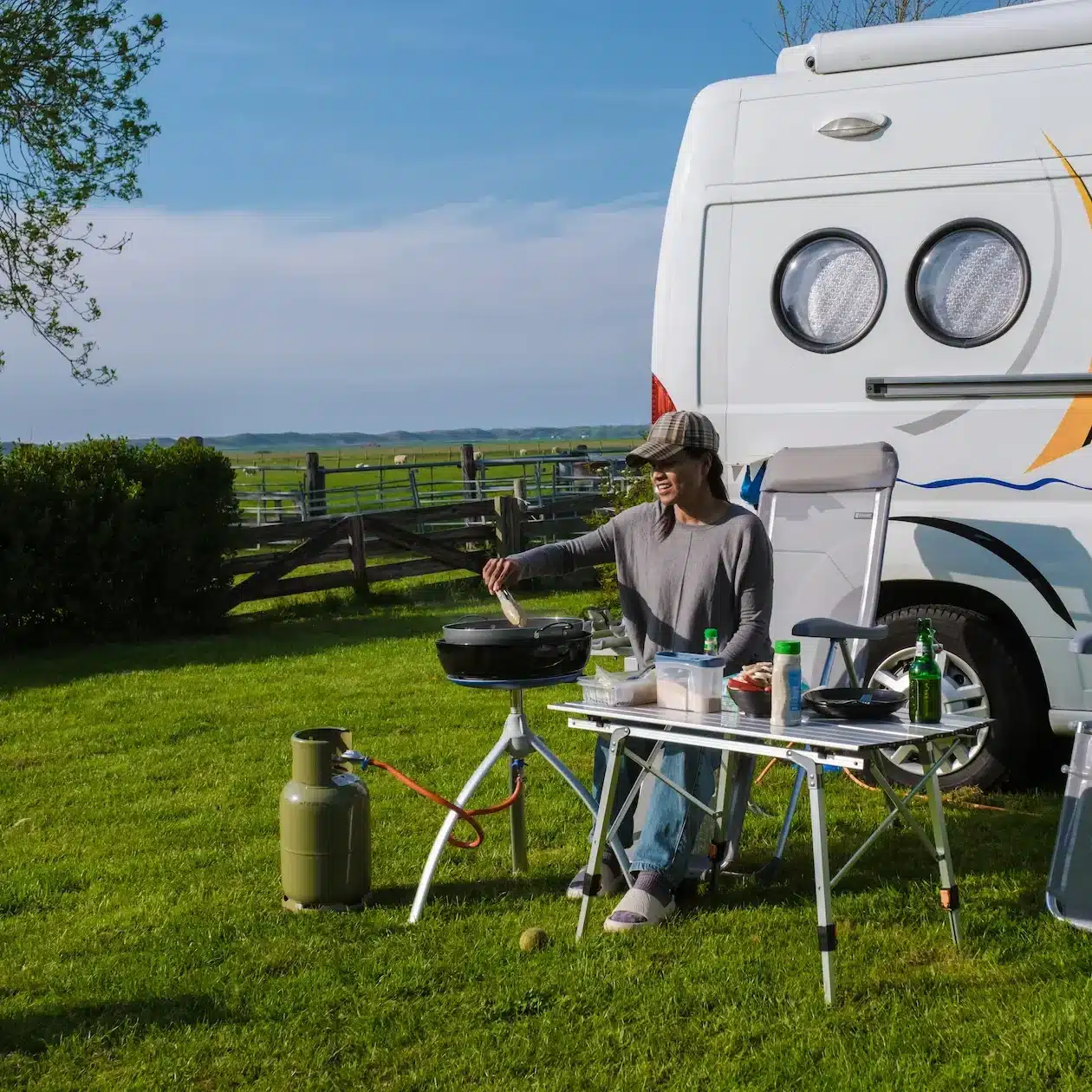 A person enjoys cooking at a portable table beside an RV, set in a lush green field with camping chairs under a clear blue sky.