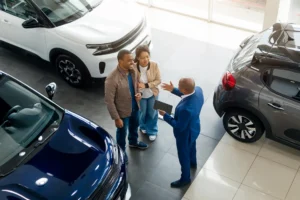 A car salesman in a blue suit is showing a tablet to a couple in a car dealership.